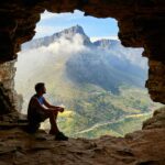 A man sitting in a cave overlooking a majestic mountain landscape under daylight.